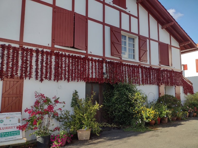 Casa encalada de blanco, con tejado a dos aguas y dos pisos. Vigas y postigos típicos franceses pintados de rojo brillante. Pimientos en la fachada, plantas en la acera a lo largo de toda la fachada principal.